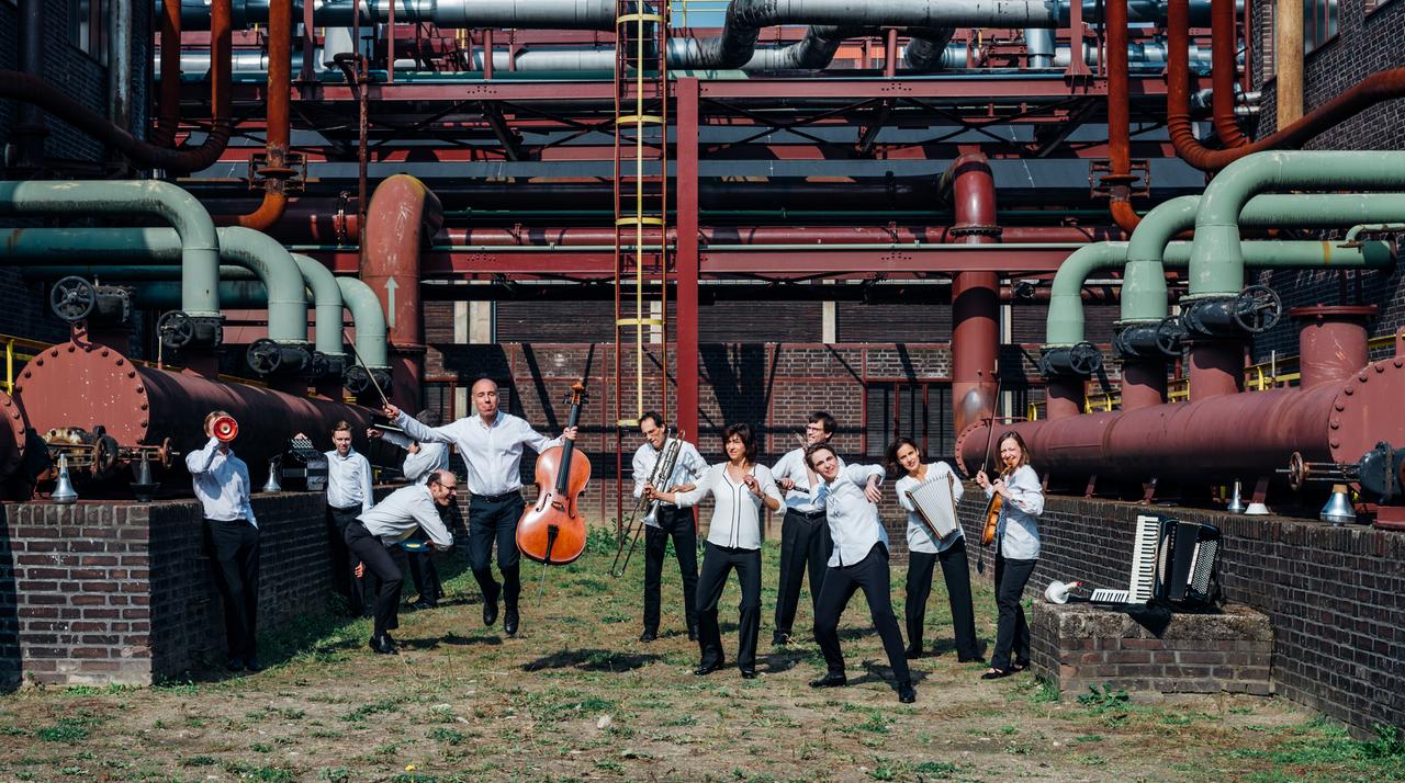 Die Mitglieder des E-MEX Ensemble feixen vor einem Industriegebäude. Einige halten ihre Instrumente in der Hand. Die Mitglieder des E-MEX Ensemble feixen vor einem Industriegebäude. Einige halten ihre Instrumente in der Hand.