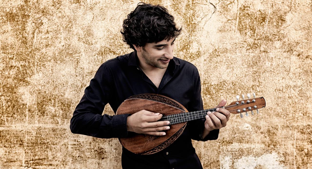 Ein Mann mit kurzen, dunklen Locken lehnt an einer braunen Wand und spielt seine aufwendig geschnitzte Mandoline. Ein Mann mit kurzen, dunklen Locken lehnt an einer braunen Wand und spielt seine aufwendig geschnitzte Mandoline.