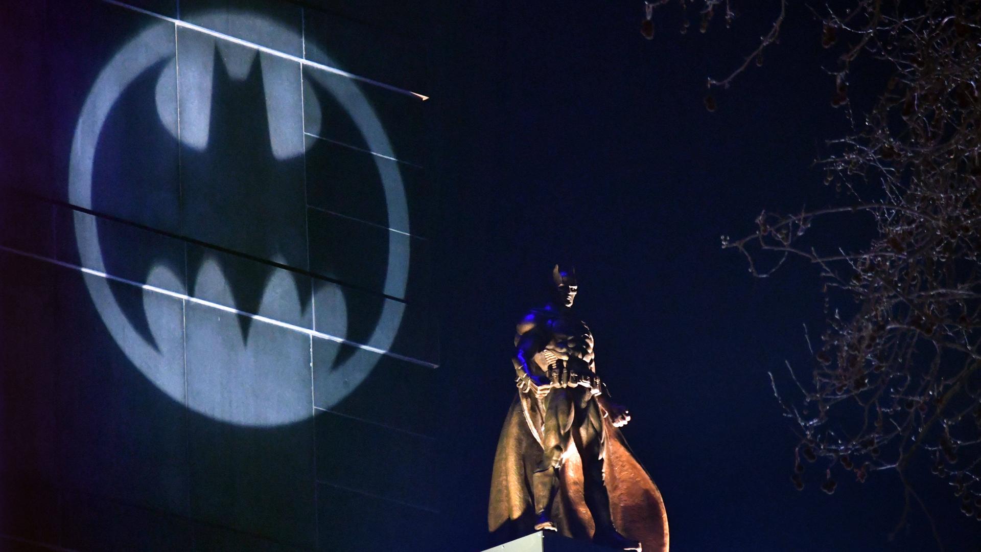 Auf einem Hochhaus wird das Symbol von Batman angestrahlt, davor steht eine große Batman-Figur auf einem anderen Häuserdach. Auf einem Hochhaus wird das Symbol von Batman angestrahlt, davor steht eine große Batman-Figur auf einem anderen Häuserdach.