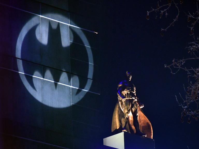 Auf einem Hochhaus wird das Symbol von Batman angestrahlt, davor steht eine große Batman-Figur auf einem anderen Häuserdach.