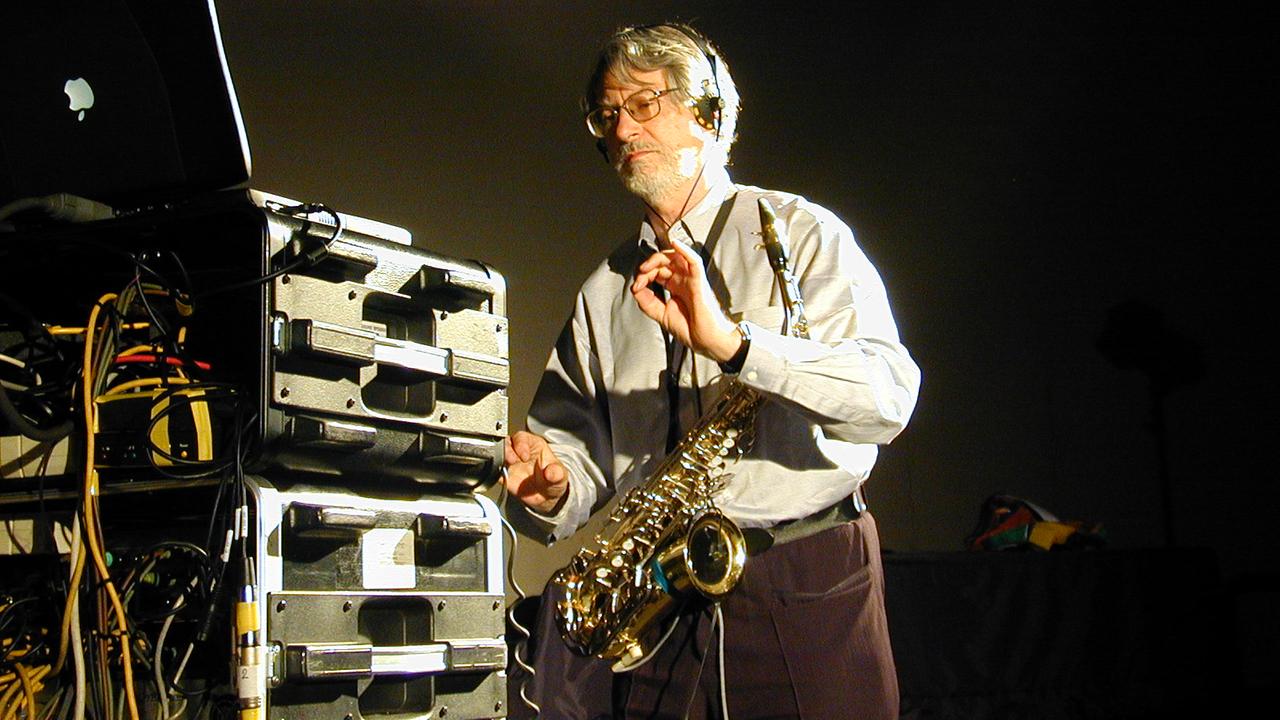 Bruno Spoerri steht mit einem Saxophon um den Hals in einem Raum, in dem Studio-Ausrüstung steht. Er trägt einen Bart und hat Kopfhörer auf, die Hände hält er wie ein Dirigent. Bruno Spoerri steht mit einem Saxophon um den Hals in einem Raum, in dem Studio-Ausrüstung steht. Er trägt einen Bart und hat Kopfhörer auf, die Hände hält er wie ein Dirigent.