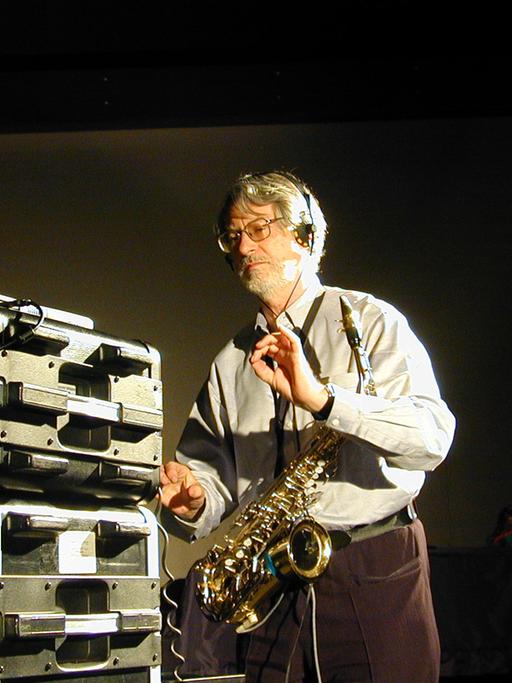 Bruno Spoerri steht mit einem Saxophon um den Hals in einem Raum, in dem Studio-Ausrüstung steht. Er trägt einen Bart und hat Kopfhörer auf, die Hände hält er wie ein Dirigent.