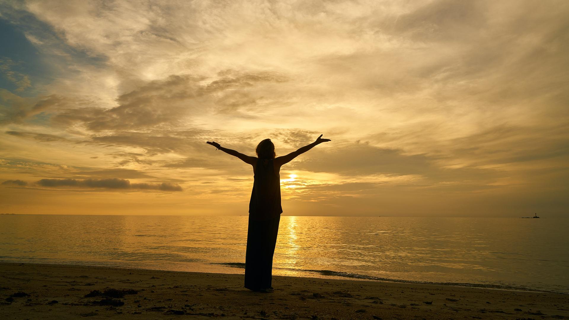 Eine weibliche Silhouette, mit zum Himmel erhobenen Armen, betrachtet den Sonnenuntergang am Meer.