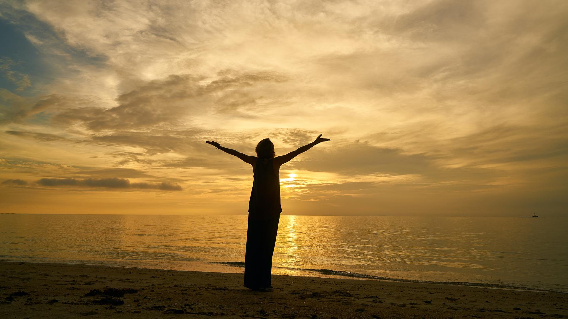 Eine weibliche Silhouette, mit zum Himmel erhobenen Armen, betrachtet den Sonnenuntergang am Meer. Eine weibliche Silhouette, mit zum Himmel erhobenen Armen, betrachtet den Sonnenuntergang am Meer.