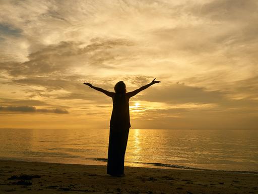 Eine weibliche Silhouette, mit zum Himmel erhobenen Armen, betrachtet den Sonnenuntergang am Meer.