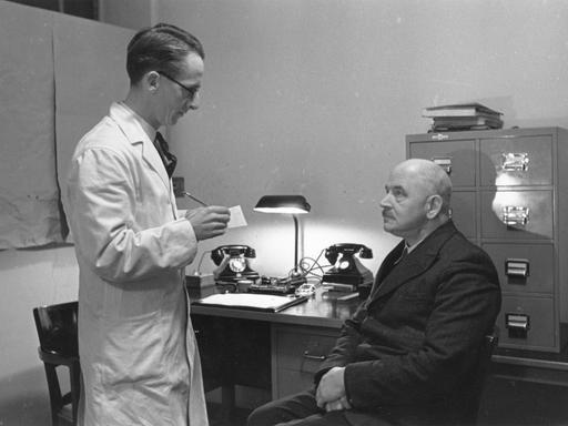 Historisches Foto einer Sprechstunde von 1941. Ein Mann sitzt auf einem Holzstuhl vor dem Allgemeinarzt im weißen Kittel.