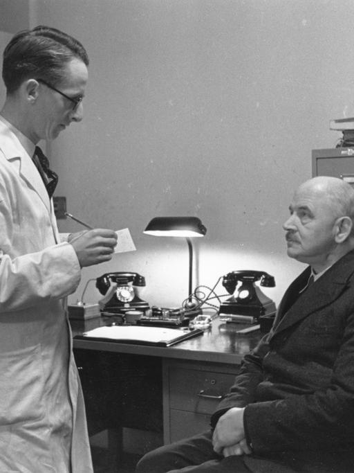 Historisches Foto einer Sprechstunde von 1941. Ein Mann sitzt auf einem Holzstuhl vor dem Allgemeinarzt im weißen Kittel.