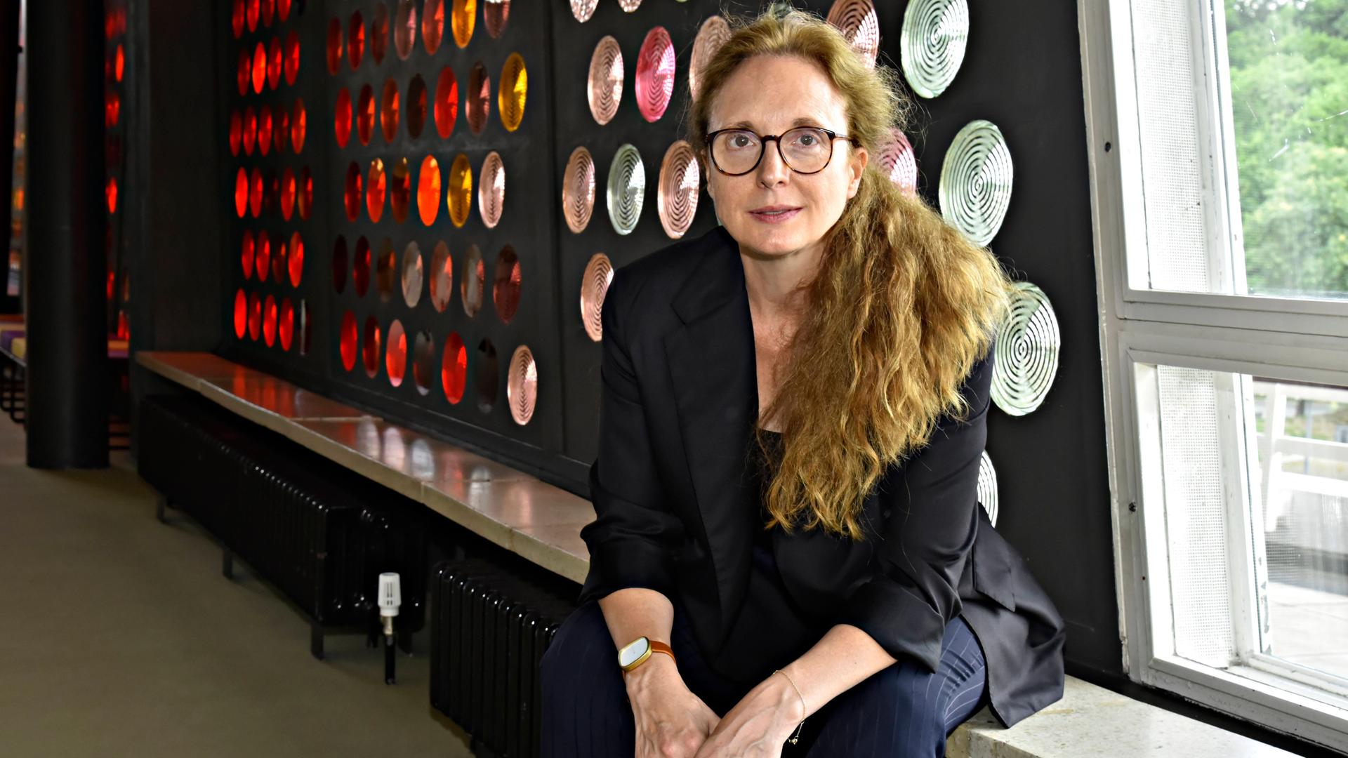 Die Komponistin Rebecca Saunders in der Philharmonie an der Herbert-von-Karajan-Straße in Berlin Tiergarten. Sie sitzt auf einer Bank über der Heizung, rechts ein Fenster, links eine Wand mit runden bunten Glasbausteinen. Die Komponistin Rebecca Saunders in der Philharmonie an der Herbert-von-Karajan-Straße in Berlin Tiergarten. Sie sitzt auf einer Bank über der Heizung, rechts ein Fenster, links eine Wand mit runden bunten Glasbausteinen.