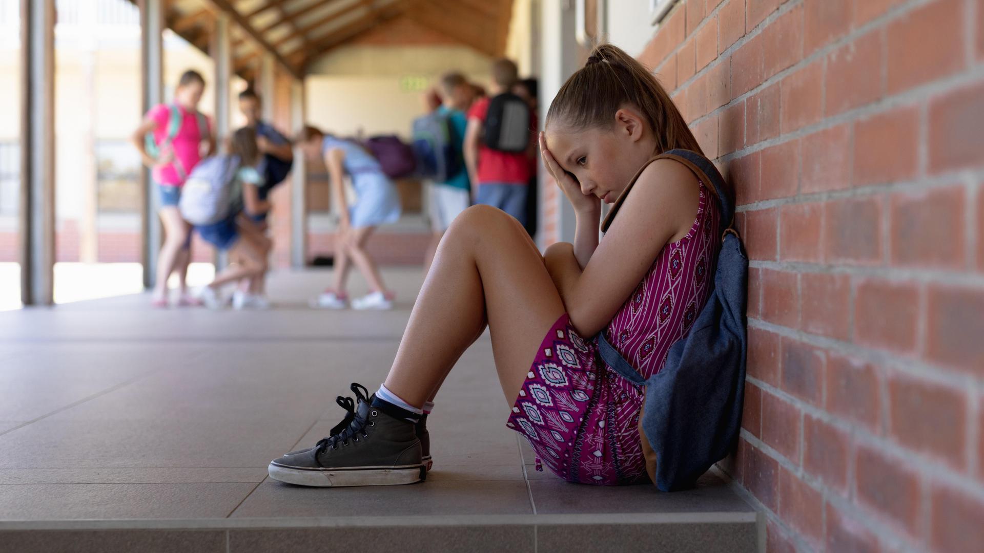 Ein blondes Mädchen im Grundschulalter, mit Turnschuhen und Rucksack, sitzt traurig allein gegen eine Wand gelehnt, während andere Kinder im Hintergrund spielen. Ein blondes Mädchen im Grundschulalter, mit Turnschuhen und Rucksack, sitzt traurig allein gegen eine Wand gelehnt, während andere Kinder im Hintergrund spielen.