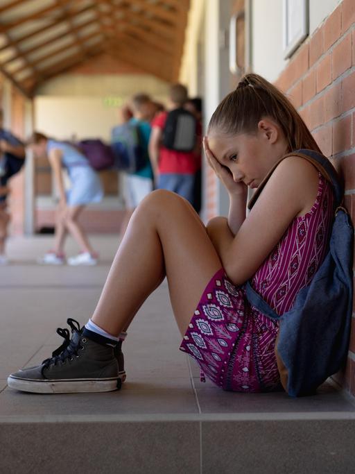 Ein blondes Mädchen im Grundschulalter, mit Turnschuhen und Rucksack, sitzt traurig allein gegen eine Wand gelehnt, während andere Kinder im Hintergrund spielen.
