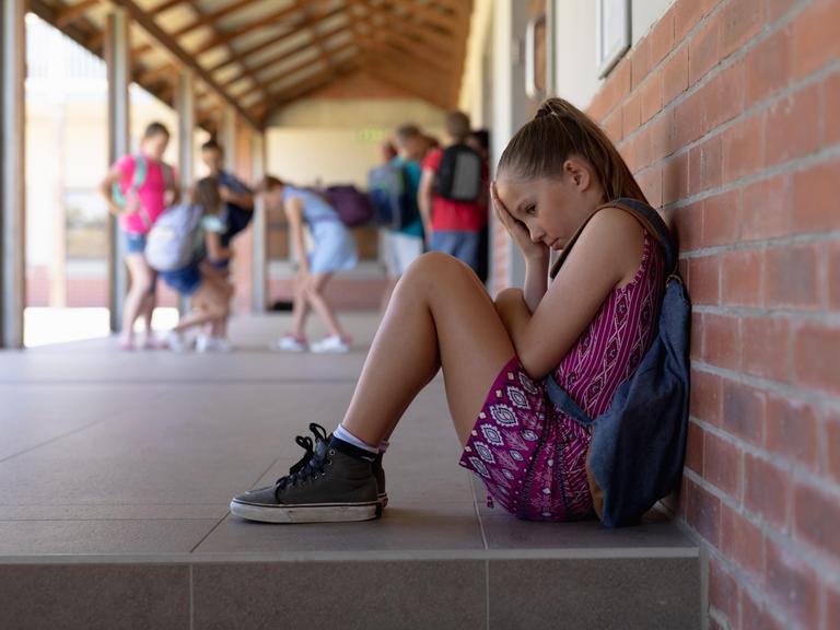 Ein blondes Mädchen im Grundschulalter, mit Turnschuhen und Rucksack, sitzt traurig allein gegen eine Wand gelehnt, während andere Kinder im Hintergrund spielen.