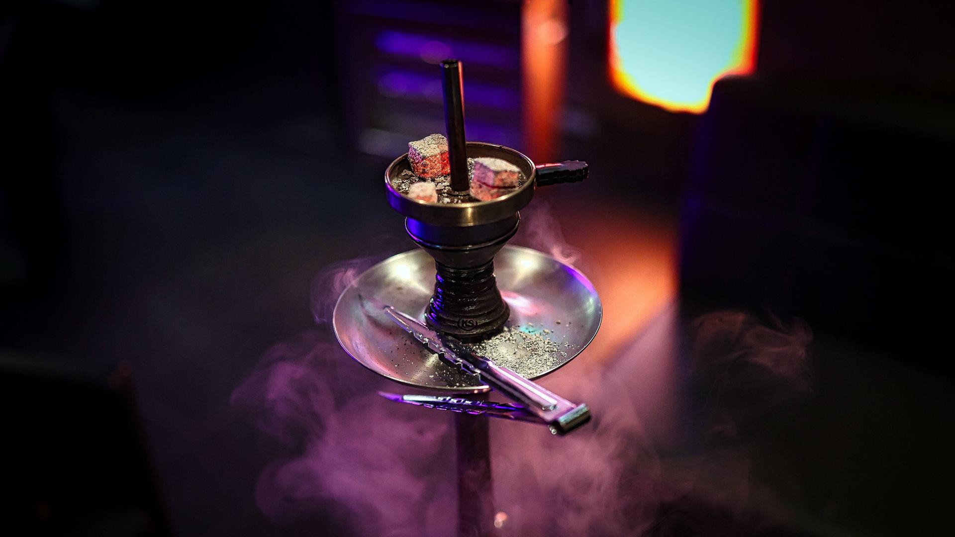 Eine Shisha Wasserpfeife mit drei glühenden Kohlen auf dem Kaminaufsatz in einer Shisha-Bar. Rauch und Neonlichter im Hintergrund. 
