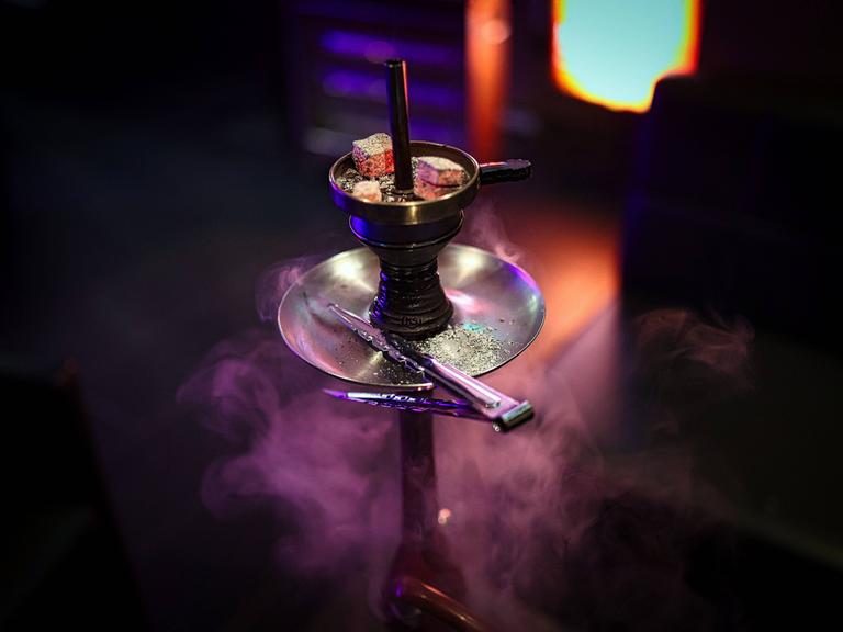 Eine Shisha Wasserpfeife mit drei glühenden Kohlen auf dem Kaminaufsatz in einer Shisha-Bar. Rauch und Neonlichter im Hintergrund. 