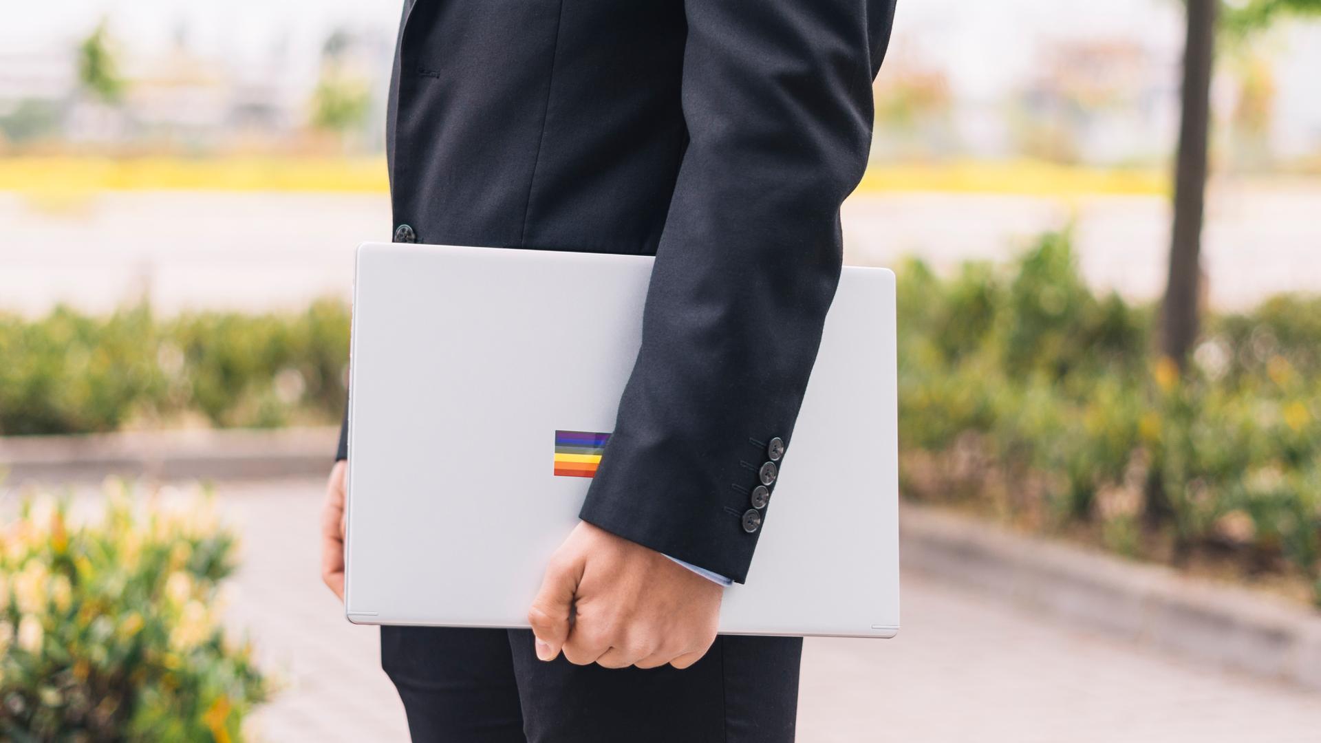 Ein Mann im Anzug hält einen Laptop unterm Arm, auf dem ein Sticker mit der Regenbogenflagge klebt. Ein Mann im Anzug hält einen Laptop unterm Arm, auf dem ein Sticker mit der Regenbogenflagge klebt.