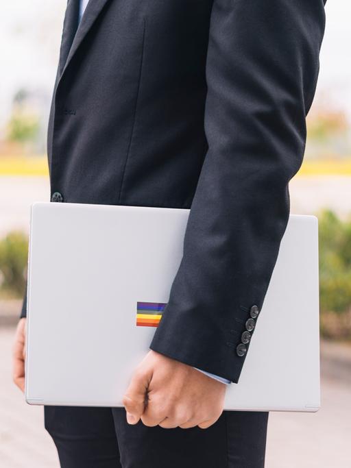 Ein Mann im Anzug hält einen Laptop unterm Arm, auf dem ein Sticker mit der Regenbogenflagge klebt.