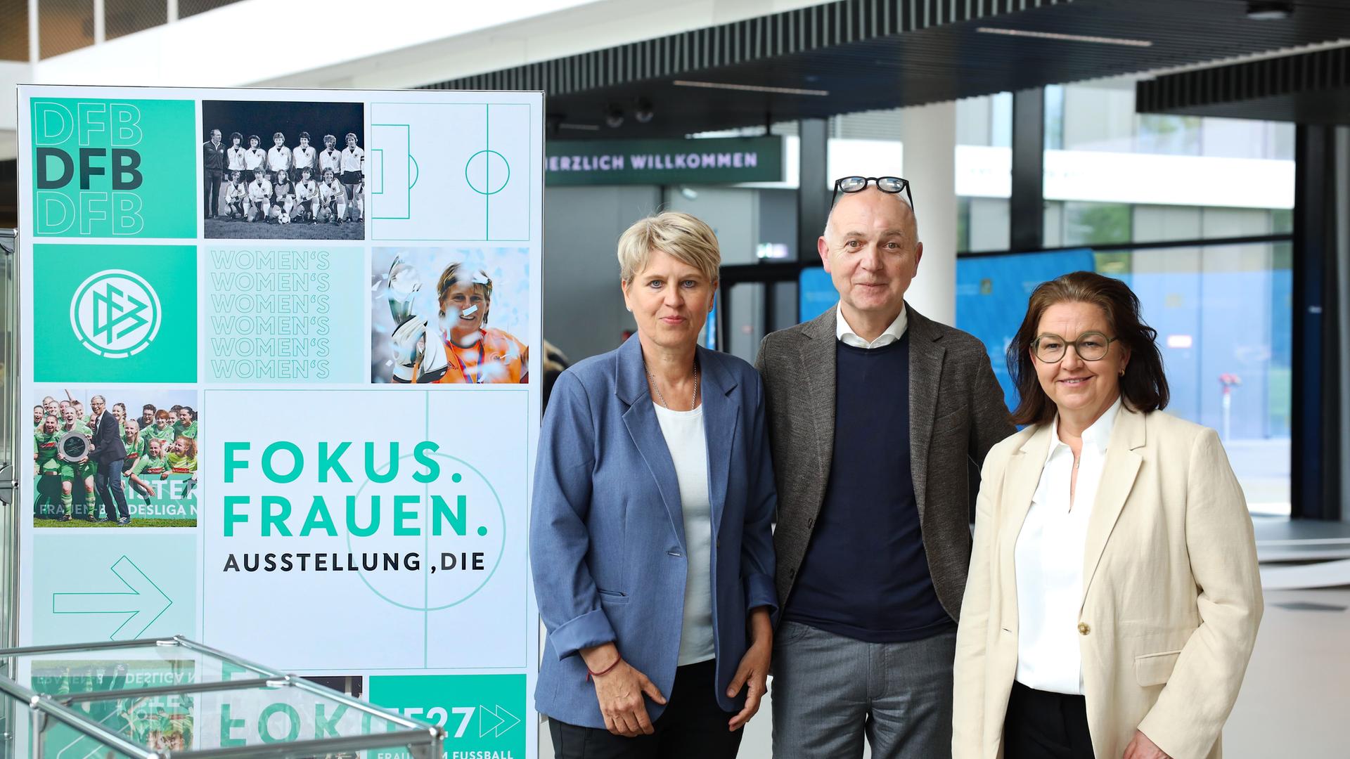 DFB Präsident Bernd Neuendorf steht zwischen Doris Fitschen, Gesamtkoordinatorin der Strategie "Frauen im Fußball" und Generalsekretärin Heike Ullrich neben einer Stellwand zu einer Ausstellung über Frauenfußball auf dem DFB Campus.
