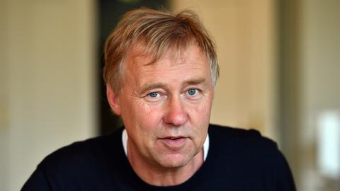 Ein Mann mit graublonden kurzen Haaren und blauen Augen blickt mit leicht geöffnetem Mund in die Kamera. Er trägt einen schwarzen Pullover.