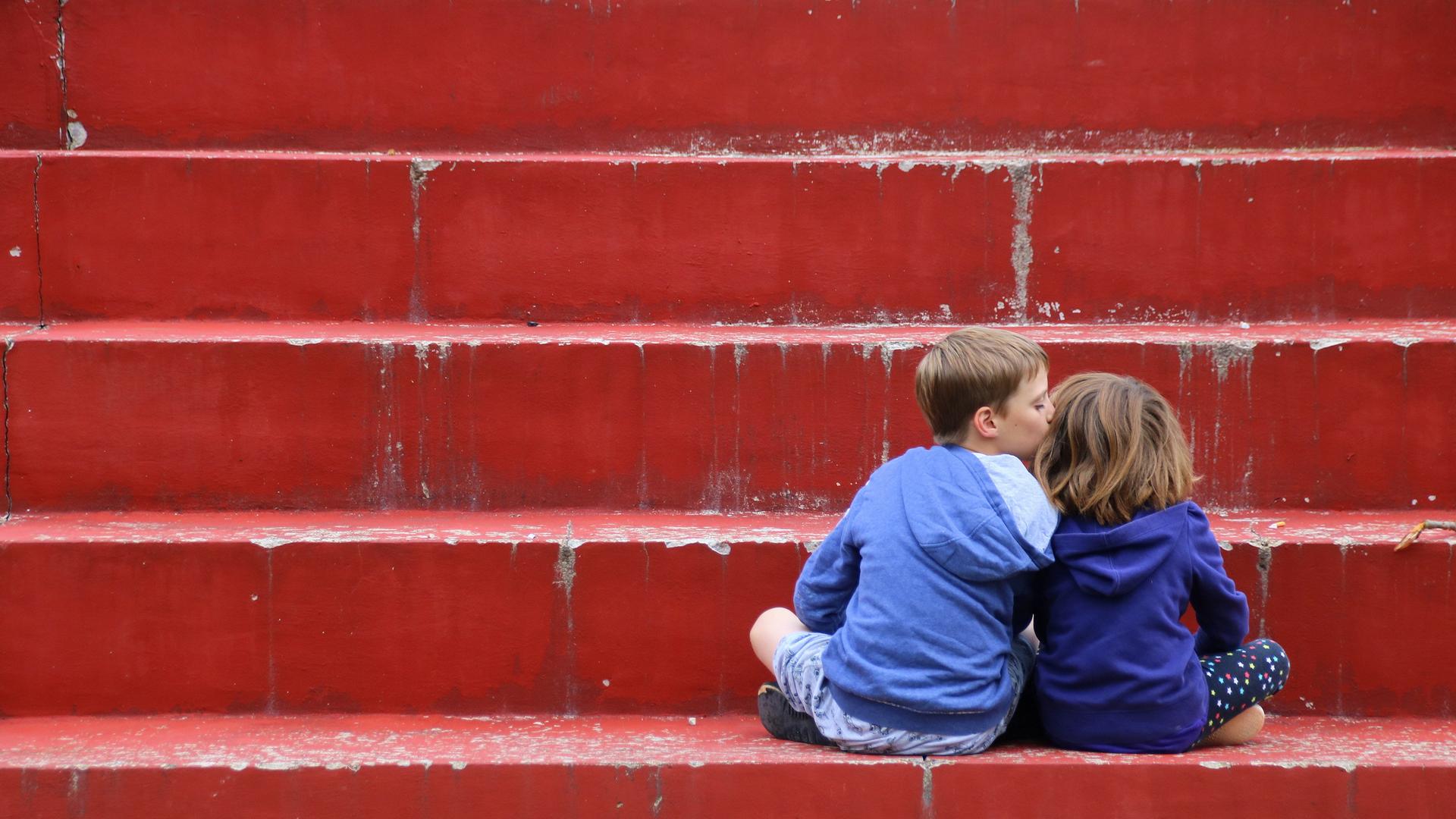 Zwei Kinder in Kapuzenpullis sitzen auf einer roten Treppe mit dem Rücken zu den Betrachtenden. Das linke Kind küsst das rechte Kind auf die Wange. Zwei Kinder in Kapuzenpullis sitzen auf einer roten Treppe mit dem Rücken zu den Betrachtenden. Das linke Kind küsst das rechte Kind auf die Wange.