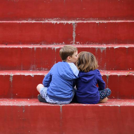 Zwei Kinder in Kapuzenpullis sitzen auf einer roten Treppe mit dem Rücken zu den Betrachtenden. Das linke Kind küsst das rechte Kind auf die Wange.