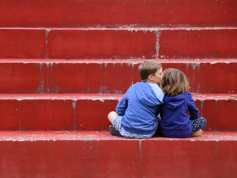 Zwei Kinder in Kapuzenpullis sitzen auf einer roten Treppe mit dem Rücken zu den Betrachtenden. Das linke Kind küsst das rechte Kind auf die Wange.