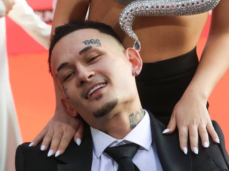 Der russische Rapper Alischer Morgenshtern sitzt lächelnd auf einem Stuhl, während er von Frauenhänden an den Schultern massiert wird.