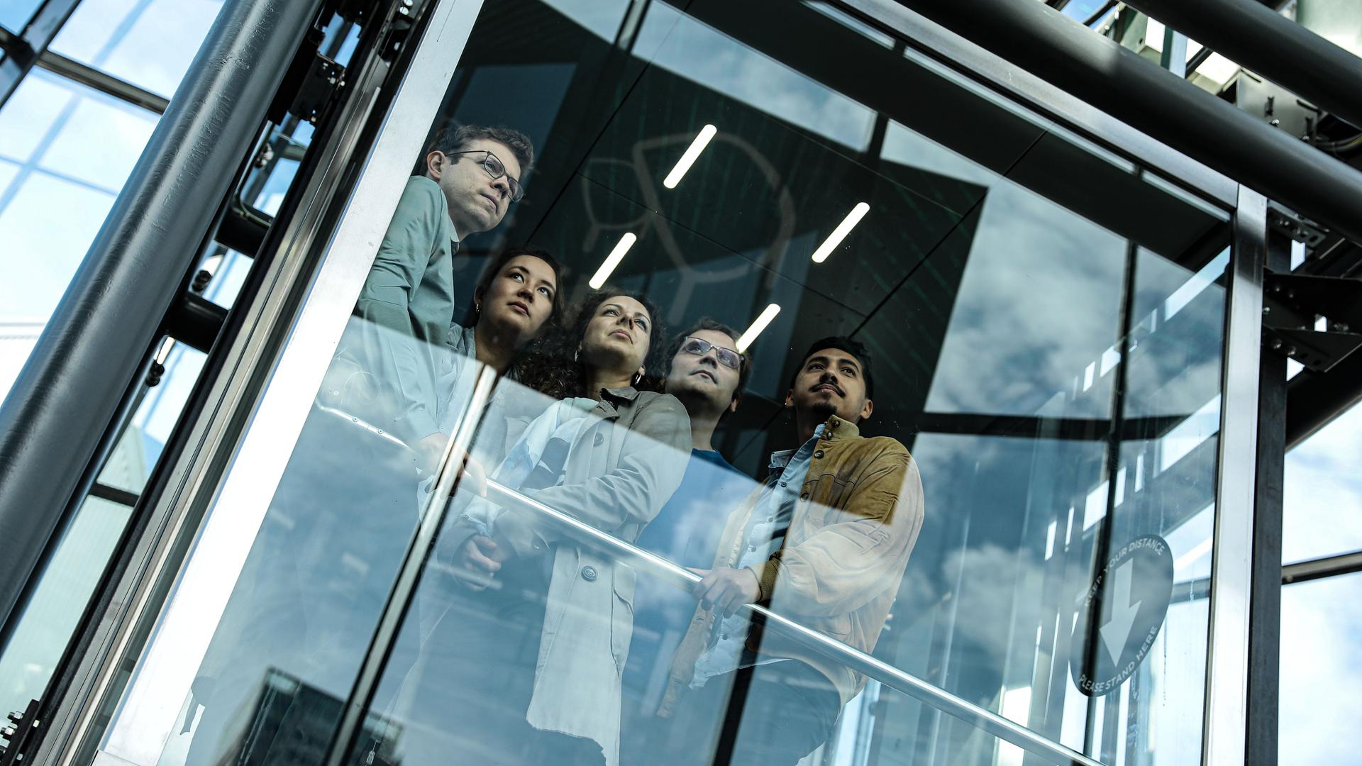 Zwei Frauen und drei Männer stehen in einem gläsernen Aufzug zusammen und schauen in den Ferne.