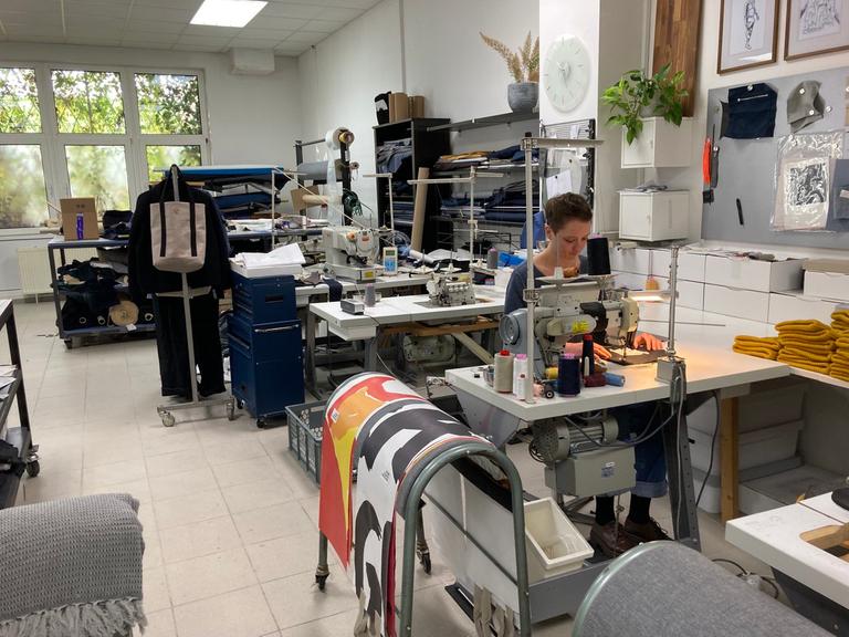 Die Schneiderin und Schnittmacherin Catharina Saffier sitzt in ihrem Berliner Atelier an einer Nähmaschine, im Hintergrund stehen weitere Maschinen und Ständer mit Stoffen und Kleidunggsstücken.
