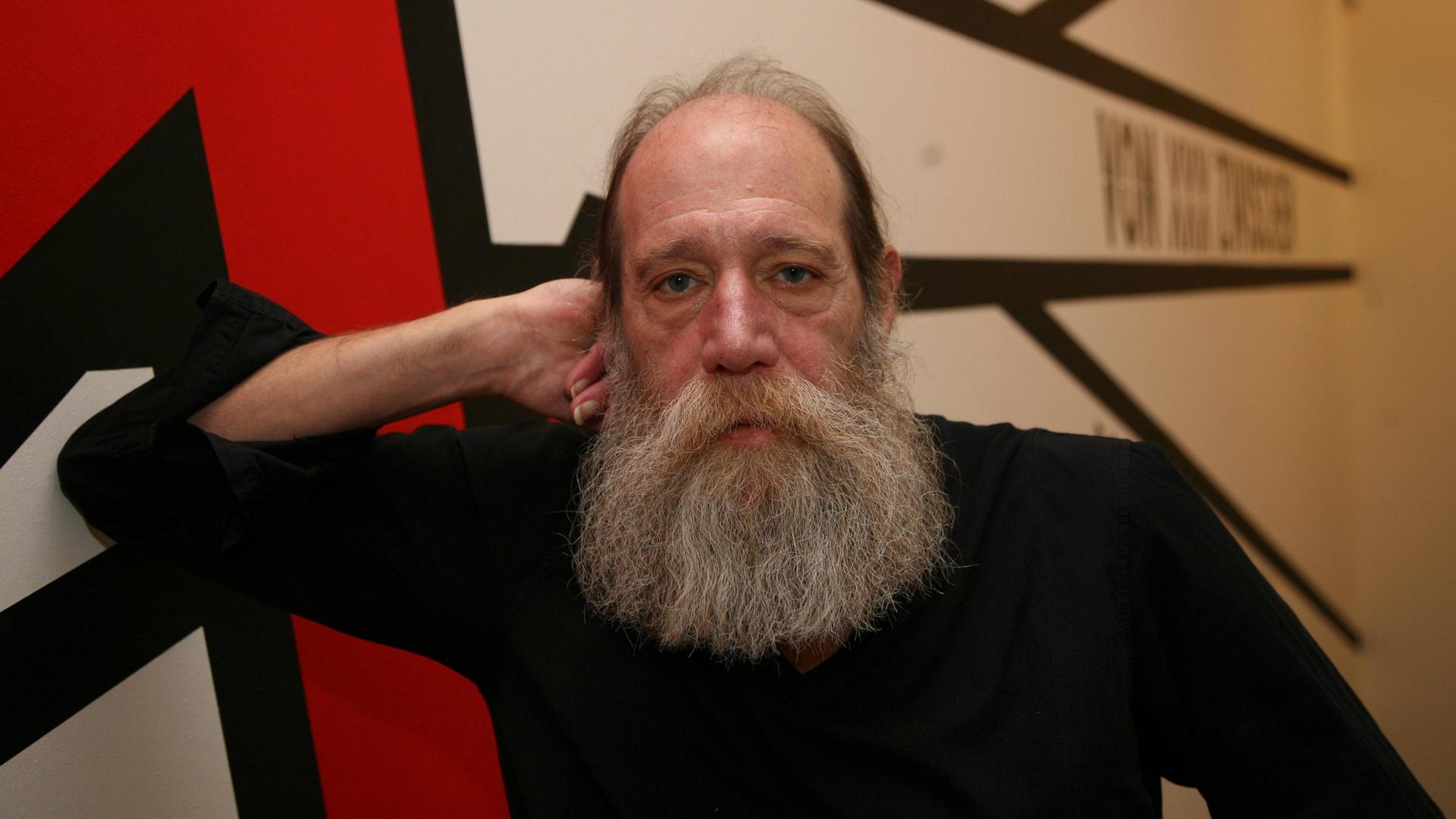 Ein Mann mit sehr langem Bart stützt seinen Kopf auf seine Faust. Hinter im sind schemenhaft große, rote Buchstaben an der Wand. Es ist Lawrence Weiner.
