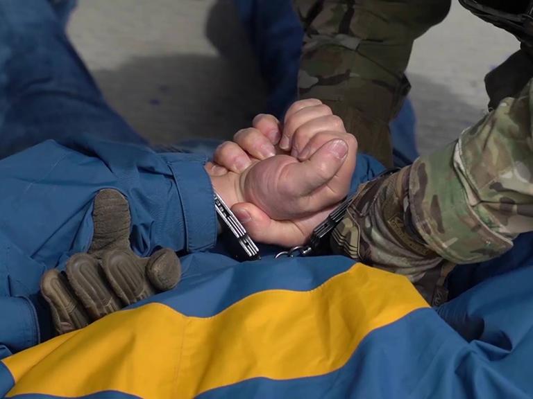 Ein Körper, der auf dem Rücken liegt, bekommt Handschellen von einer Person in einem Camouflage-Anzug angelegt