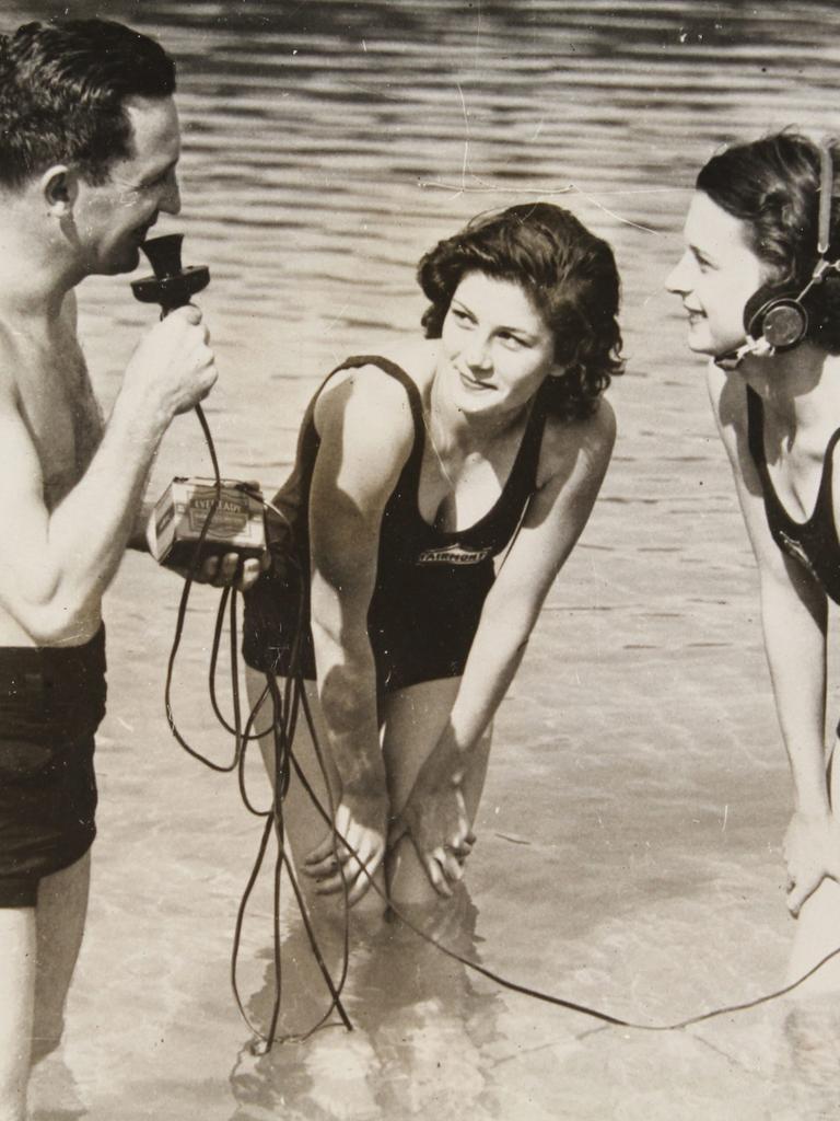 Auf dem Schwarzweißfoto von April 1934 ist links ein Mann in Badehose zu sehen, der ein Mikrofon hält und knietief im Wasser steht. Rechts von ihm stehen zwei junge Frauen, von denen eine einen Kopfhörer trägt, der über Kabel und ein Kästchen mit dem Mikro des Mannes verbunden ist. Die Frauen stehen auch im Wasser und schaeun den Mann an. 