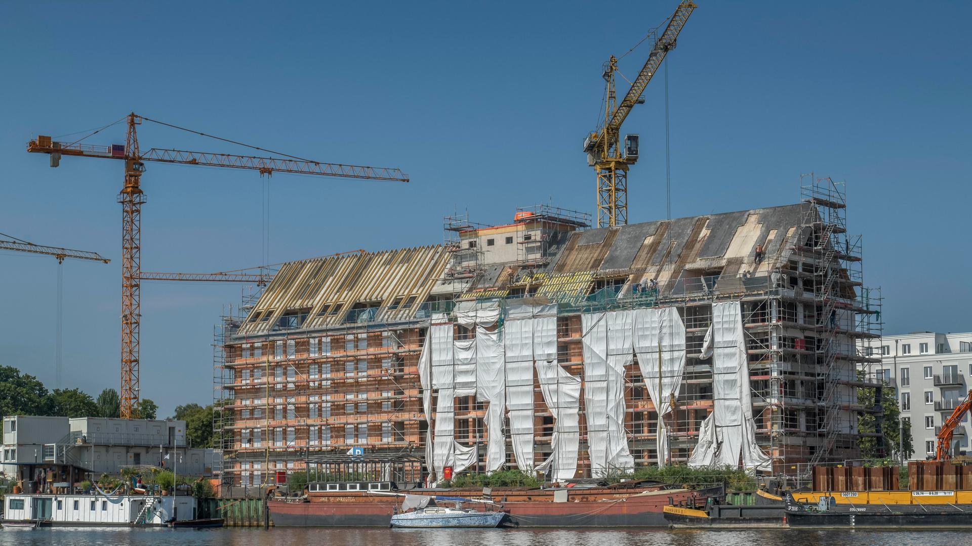 Kräne überragen ein großes eingerüstetes Gebäude, das direkt am Wasser steht. Kräne überragen ein großes eingerüstetes Gebäude, das direkt am Wasser steht.