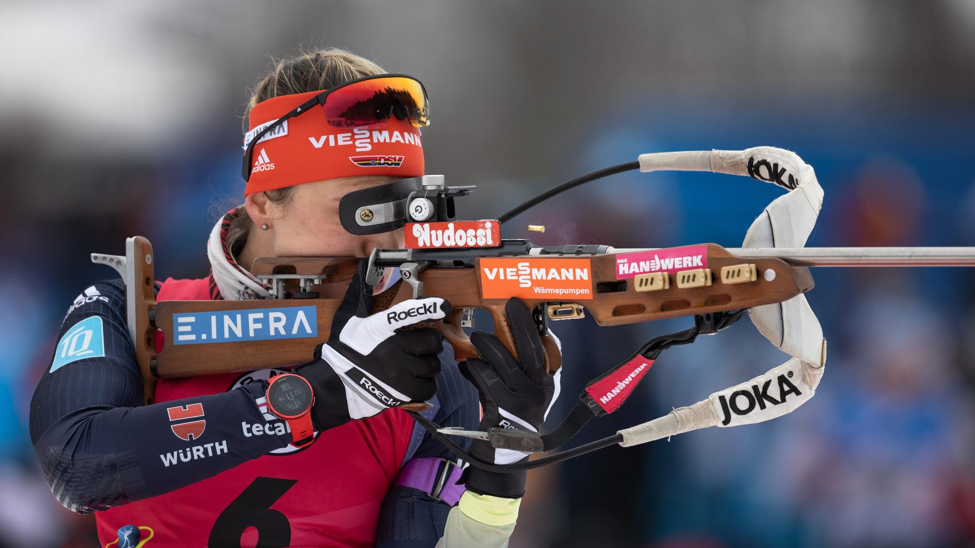 Eine Biathletin ist von der Seite zu sehen, wie sie in ihrem schwarz-roten Wettkampfanzug das Gewehr anlegt. Ihr Gewehr, das Stirnband und der Anzug der Athletin sind übersät mit den Logos ihrer Sponsoren. Eine Biathletin ist von der Seite zu sehen, wie sie in ihrem schwarz-roten Wettkampfanzug das Gewehr anlegt. Ihr Gewehr, das Stirnband und der Anzug der Athletin sind übersät mit den Logos ihrer Sponsoren.