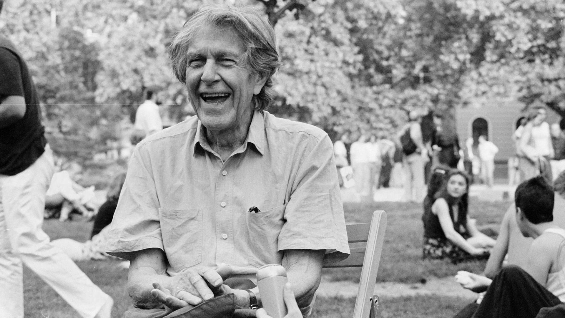John Cage sitzt lachend auf einer Wiese und gibt ein Interview in ein Reportermikrofon, im Hintergrund sitzen und gehen junge Leute in Freizeitkleidung. John Cage sitzt lachend auf einer Wiese und gibt ein Interview in ein Reportermikrofon, im Hintergrund sitzen und gehen junge Leute in Freizeitkleidung.