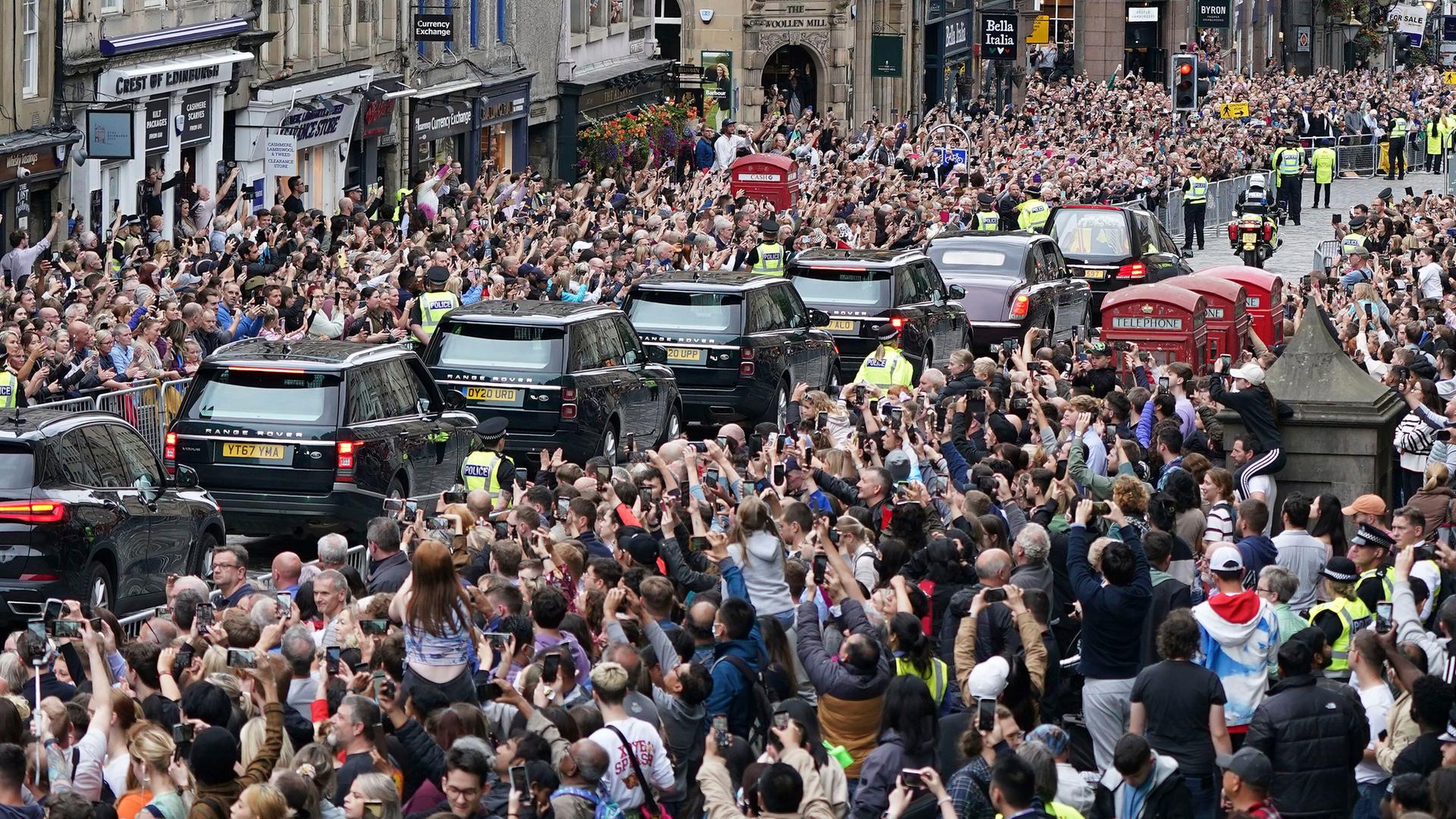In Edinburgh stehen tausende Menschen am Straßen-Rand. Sie sehen Autos mit dem Sarg von Königin Elisabeth vorbei fahren. In Edinburgh stehen tausende Menschen am Straßen-Rand. Sie sehen Autos mit dem Sarg von Königin Elisabeth vorbei fahren.