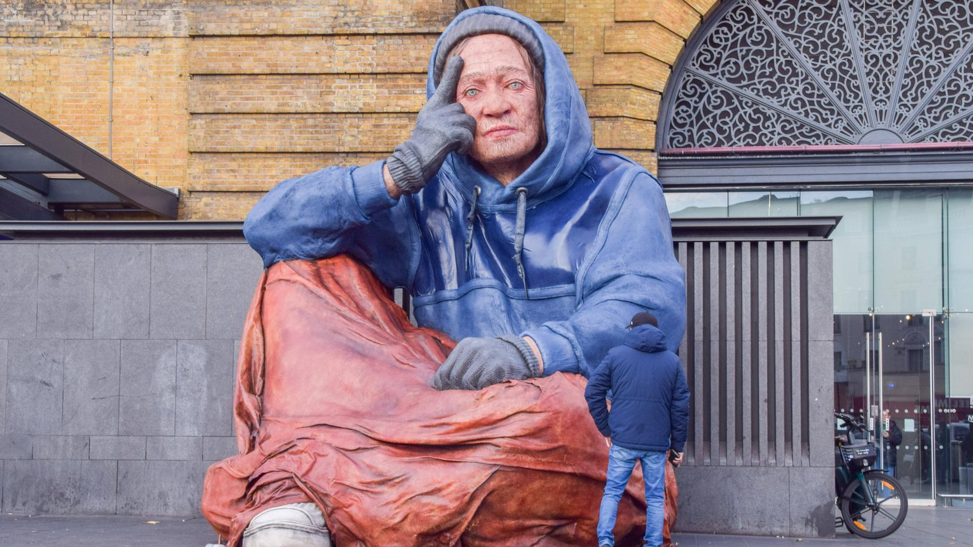 Vor der überlebensgroßen Skulptur einer obdachlosen Person steht ein Mann im Anorak, der der sitzenden Figur gerade bis zum Ellenbogen reicht.