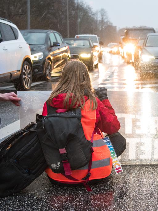 Zwei junge Frauen in orangen Warnwesten mit einem Transparent knien auf einer Fahrbahn, um sie zu blockieren. Ihnen gegenüber: PKWs mit leuchtenden Scheinwerfern. 