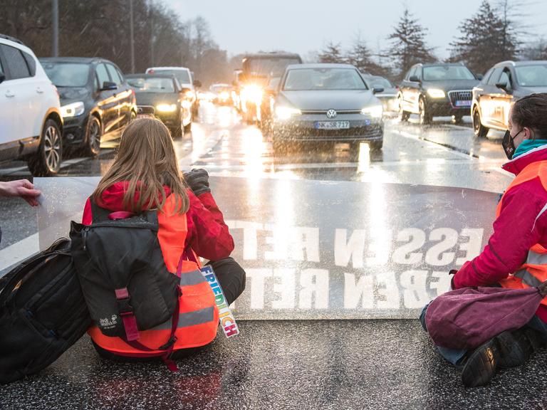 Zwei junge Frauen in orangen Warnwesten mit einem Transparent knien auf einer Fahrbahn, um sie zu blockieren. Ihnen gegenüber: PKWs mit leuchtenden Scheinwerfern. 