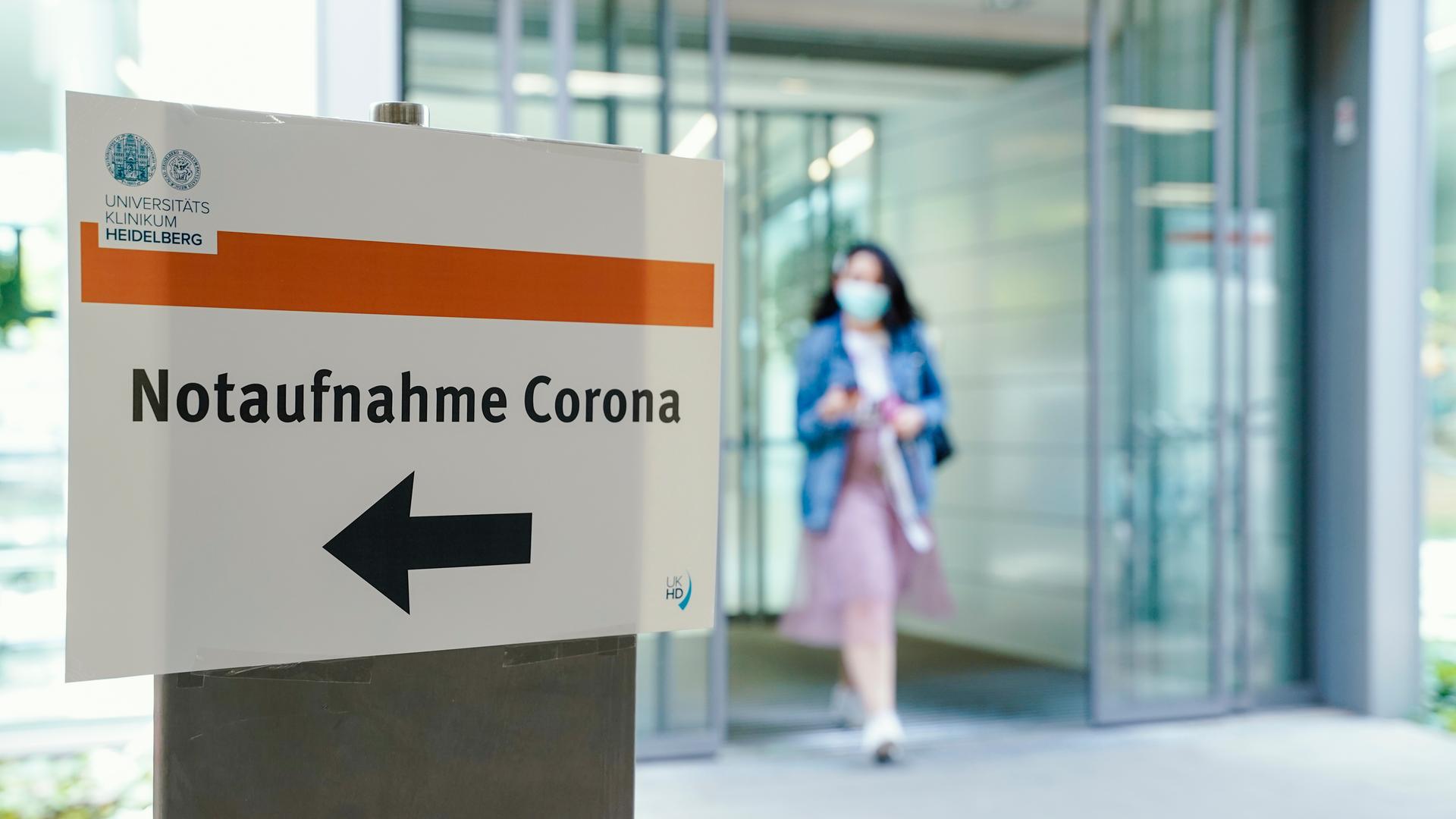 Eine Frau kommt aus einem Krankenhaus, im Vordergrund ist ein Schild mit der Aufschrift "Notaufnahme Corona" zu sehen Eine Frau kommt aus einem Krankenhaus, im Vordergrund ist ein Schild mit der Aufschrift "Notaufnahme Corona" zu sehen