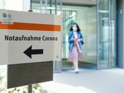 Eine Frau kommt aus einem Krankenhaus, im Vordergrund ist ein Schild mit der Aufschrift "Notaufnahme Corona" zu sehen Eine Frau kommt aus einem Krankenhaus, im Vordergrund ist ein Schild mit der Aufschrift "Notaufnahme Corona" zu sehen