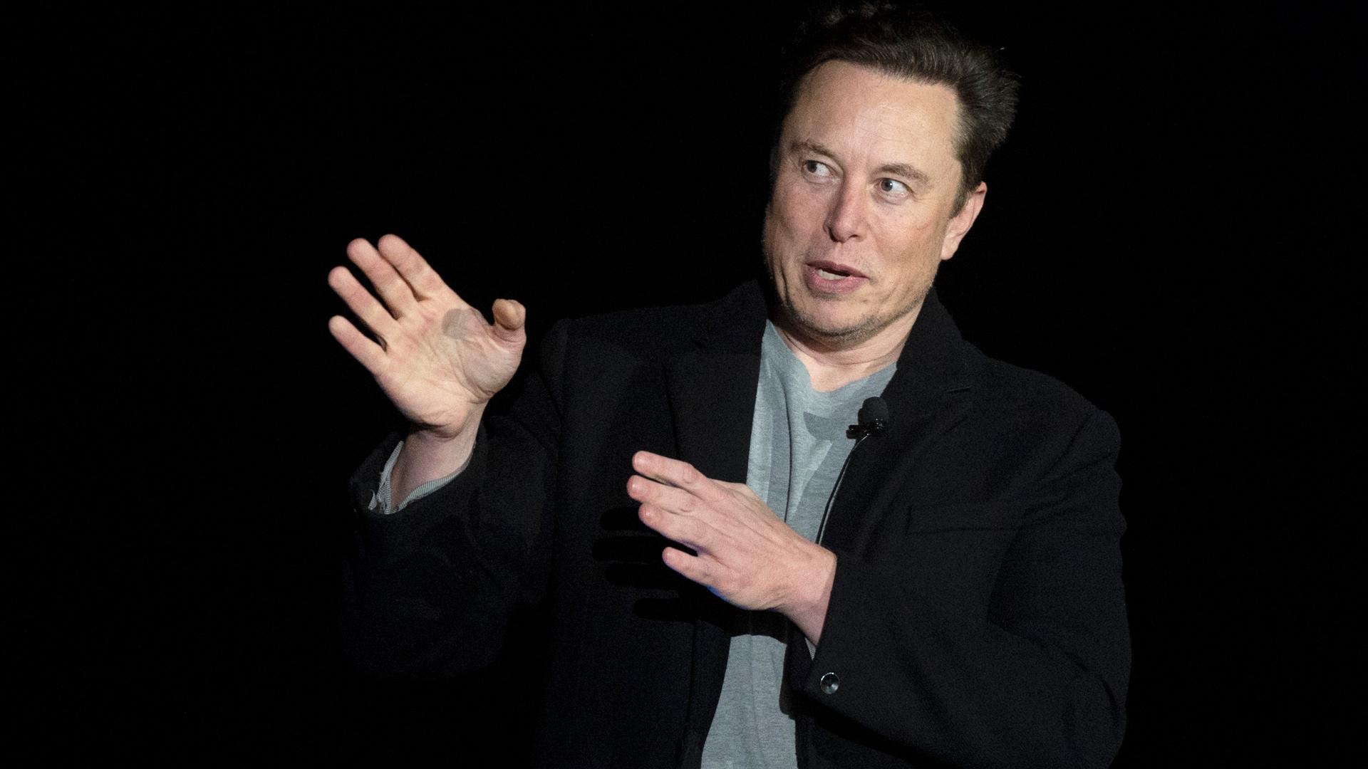 Der Tech-Unternehmer Elon Musk macht während eines Vortrags mit beiden Händen eine Geste, als wollte er etwas beiseite schieben.