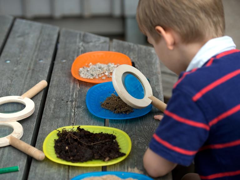 Kinder der Kita Josephinenstraße in Dresden (Sachsen) betrachten mit einem Vergrößerungsglas auf bunten Tellern Erde, Sand und Kieselsteine.