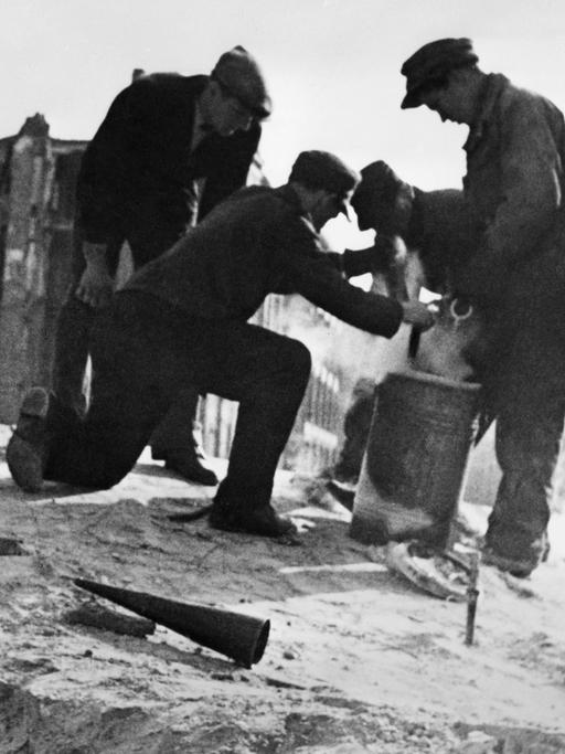 Das Schwarz-Weiß-Foto zeigt drei Männer vor beschädigten Fassaden im Jahr 1947 in der Berliner Mauerstraße. Sie hantieren mit Metalleimern und bereiten die Sprengung des Bunkers der Gauleitung der NSDAP vor. Er war in die Ruine der Dreifaltigkeitskirche eingebaut worden.