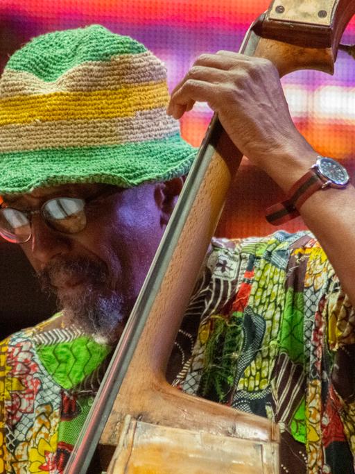 Ein Mann in grellbuntem Hemd und ebenso bunter Strickhut spielt Kontrabass. Er trägt Brille und hat einen Bart.