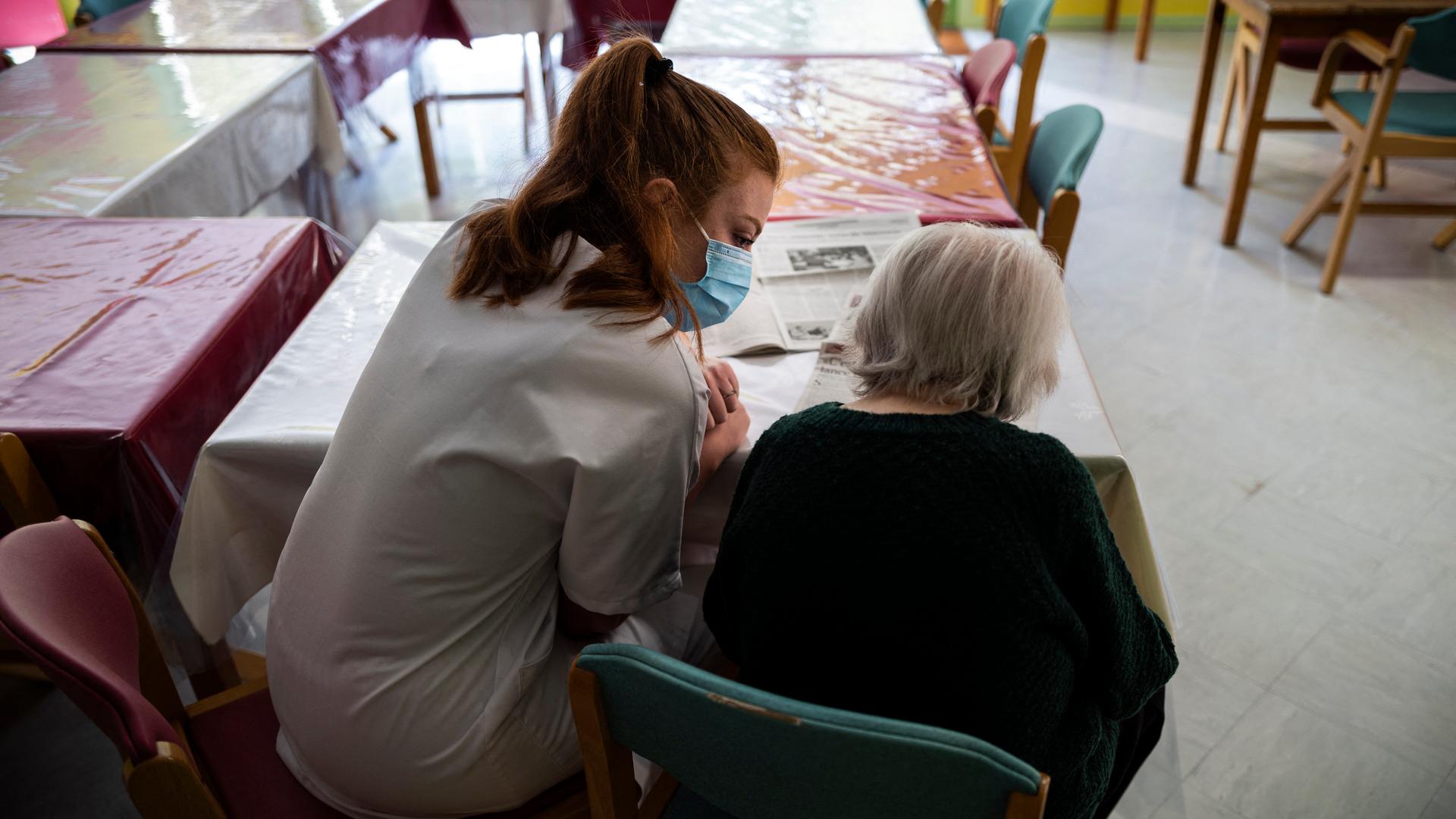 Eine weiß gekleide Krankenschwester sitzt mit einer alten Frau am Tisch und liest gemeinsam mit ihr die Zeitung. Eine weiß gekleide Krankenschwester sitzt mit einer alten Frau am Tisch und liest gemeinsam mit ihr die Zeitung.