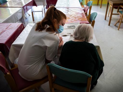Eine weiß gekleide Krankenschwester sitzt mit einer alten Frau am Tisch und liest gemeinsam mit ihr die Zeitung.