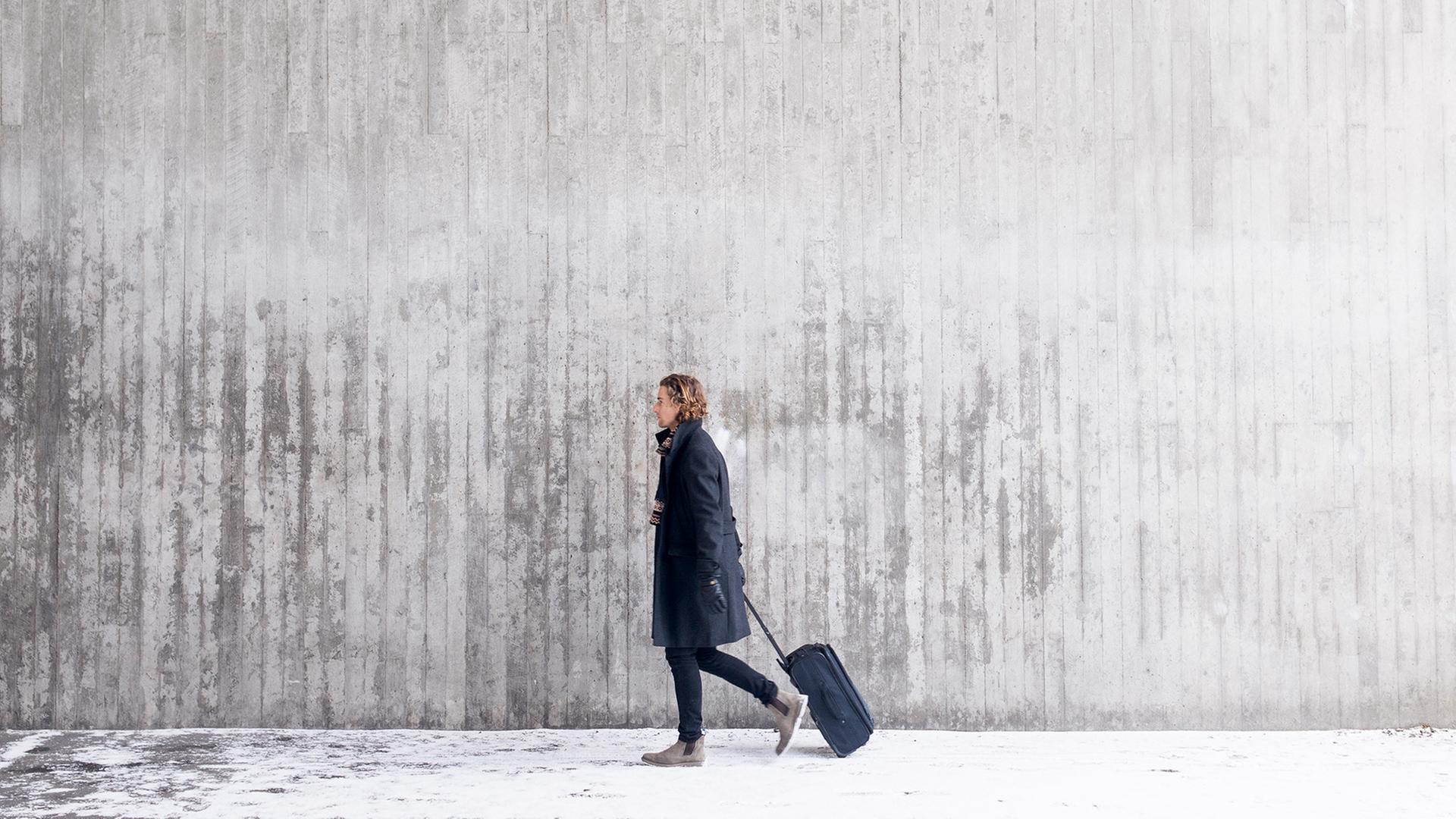 Eine Frau vor einer grauen Wand zieht einen Koffer hinter sich her. 