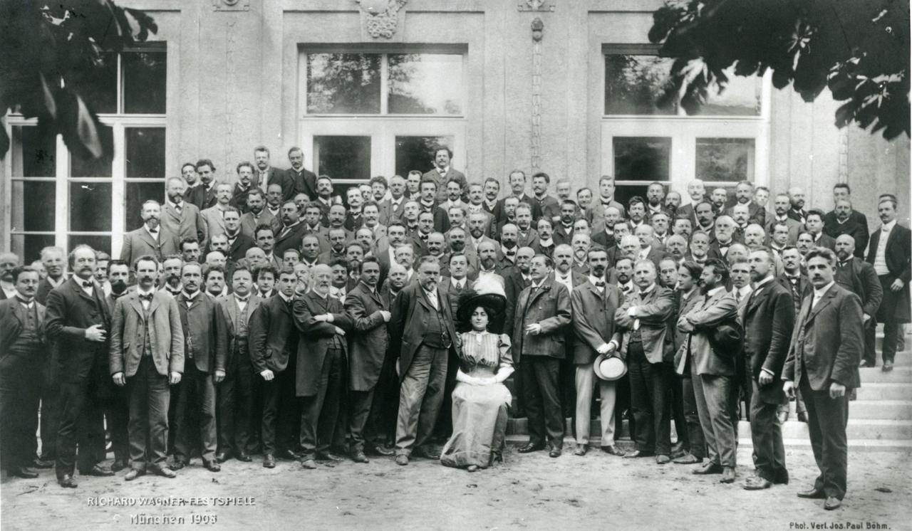 Schwarz-weiß-Aufnahme von 1908 von Orchestermitgliedern, die ohne Instrumente mit einer Sängerin für ein Foto posieren. Sie stehen draußen vorder Wand eines großen Gebäudes.