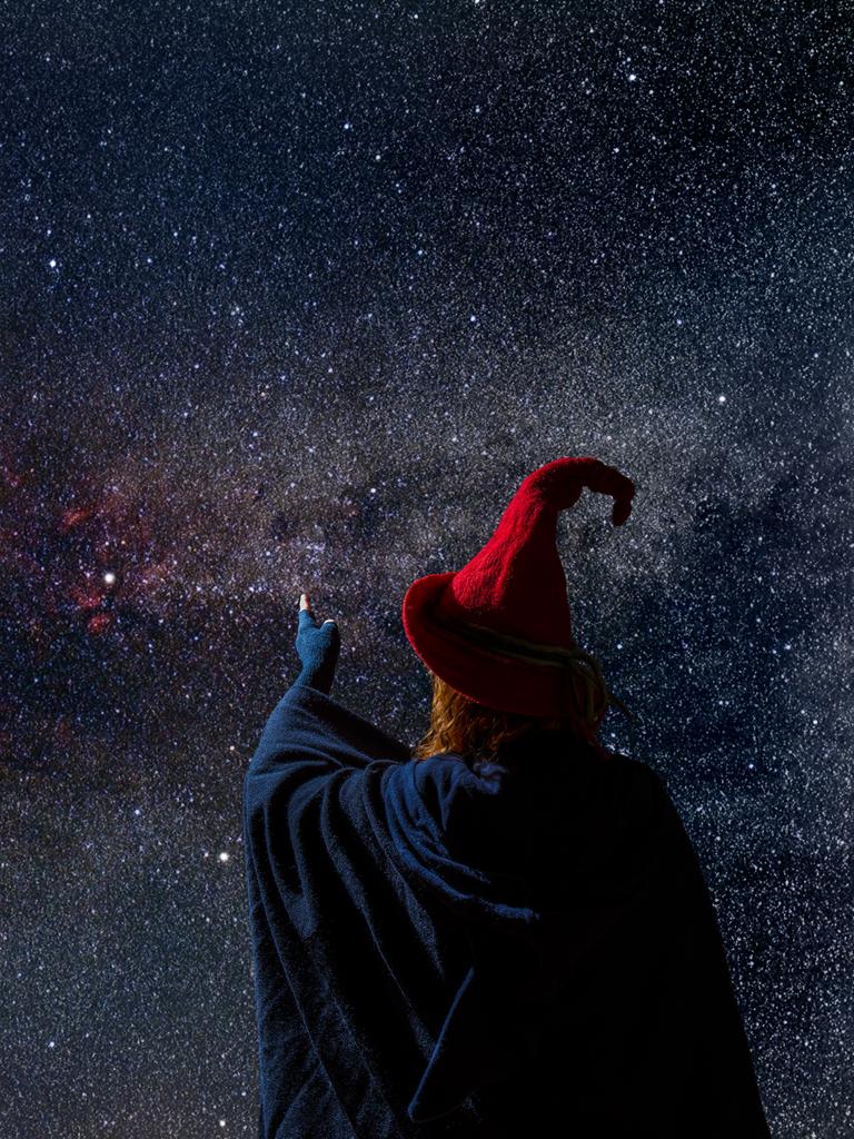 Eine Person mit blauem Umhang, rotem Spitzhut und fingerlosen Handschuhen steht vor einem nächtlichen Sternenhimmel.