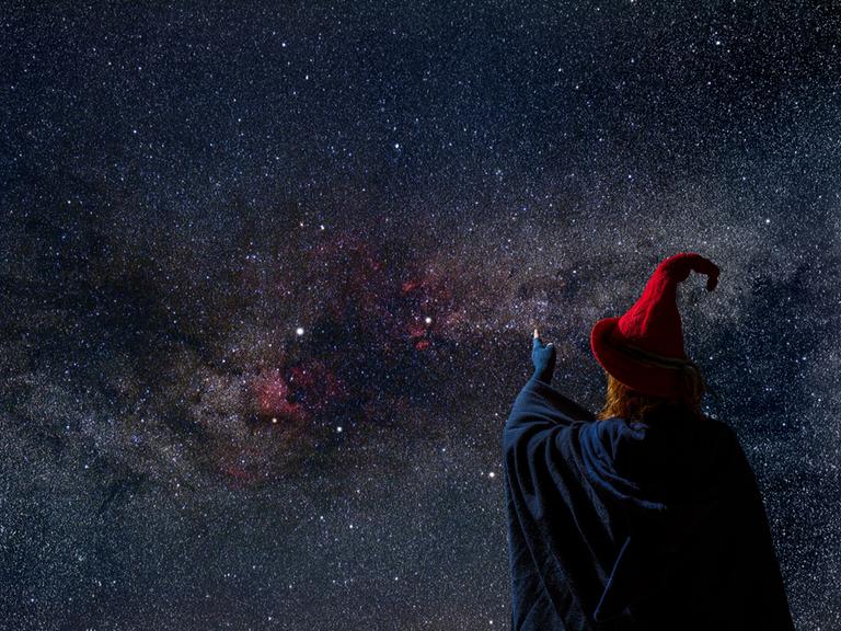 Eine Person mit blauem Umhang, rotem Spitzhut und fingerlosen Handschuhen steht vor einem nächtlichen Sternenhimmel.