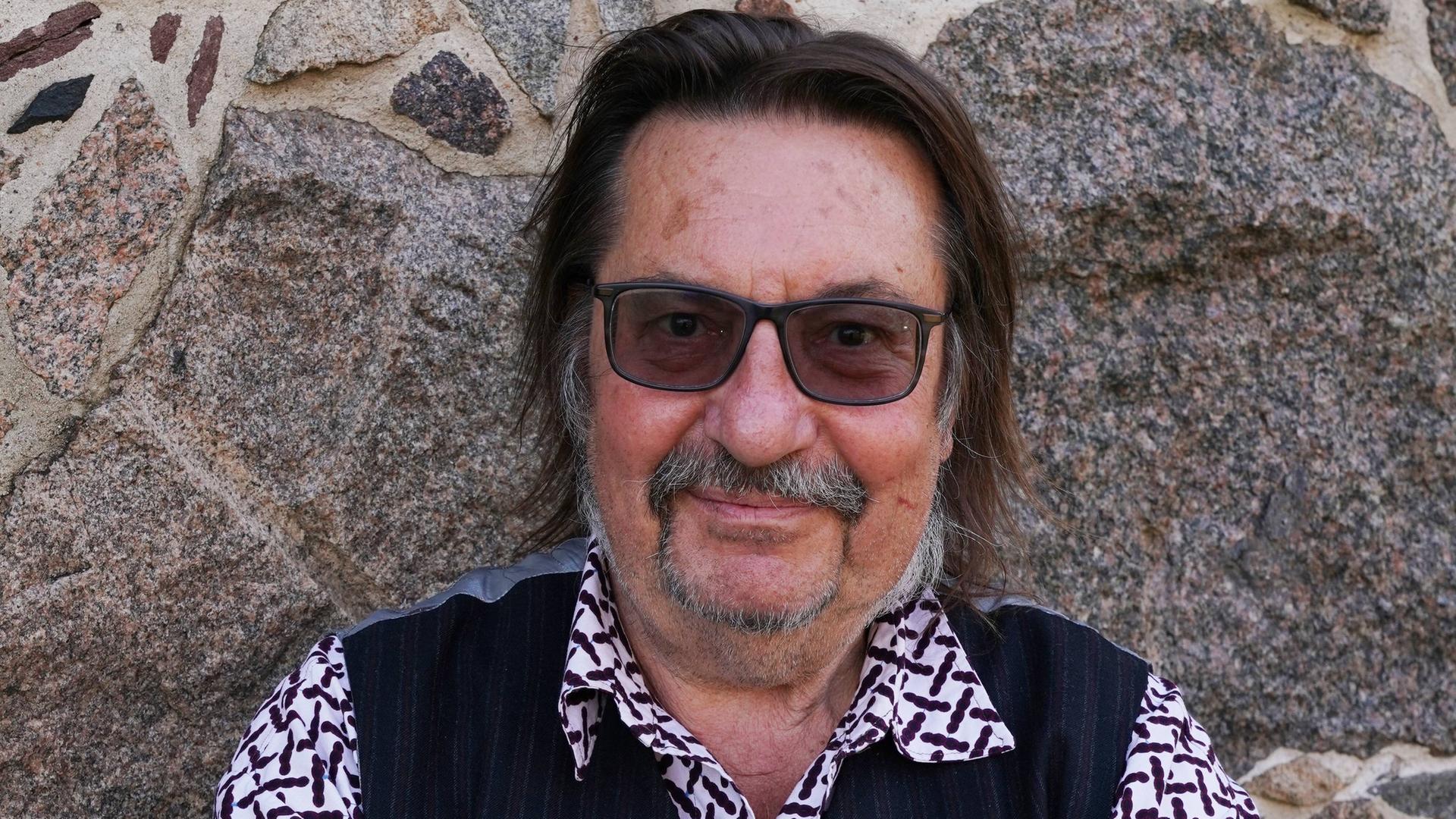 Ein Mann mit einer getönten Brille steht vor einer Steinwand. Er trägt kariertes Hemd und eine dunkle Weste. Er hält die Arme verschränkt und blickt freundlich. Es ist der Musiker Manfred Maurenbrecher. Ein Mann mit einer getönten Brille steht vor einer Steinwand. Er trägt kariertes Hemd und eine dunkle Weste. Er hält die Arme verschränkt und blickt freundlich. Es ist der Musiker Manfred Maurenbrecher.